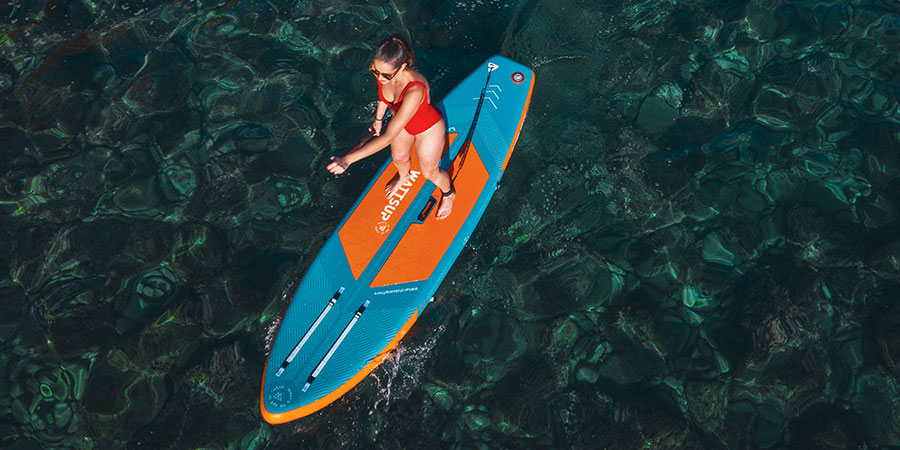 Jeune femme en maillot de bain rouge debout sur la planche de stand up paddle Wattsup Lined 10’2, pagayant dans une eau bleu turquoise foncé.